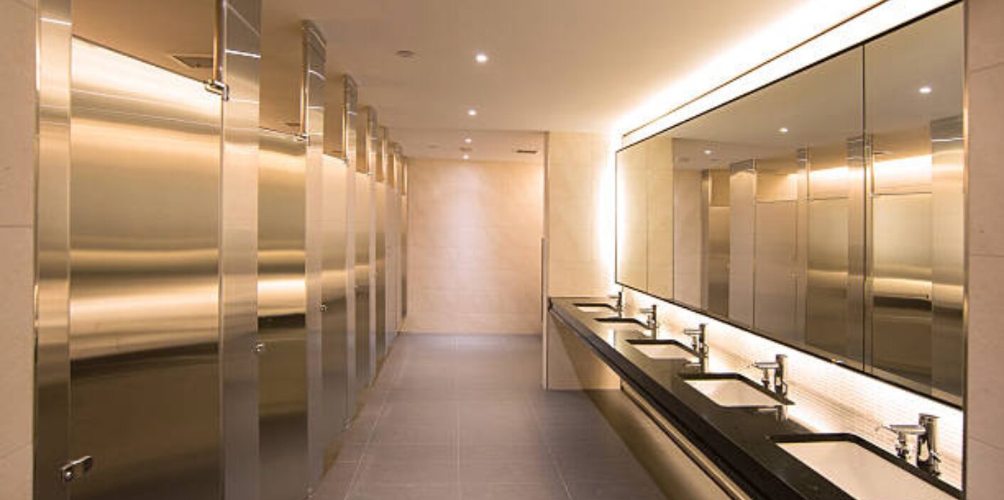 A long hallway of modern, stainless steel restroom stalls and sinks in an office building, representing the need for reliable commercial plumbing.