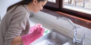 A homeowner wearing pink gloves cleans a kitchen sink to prevent clogged drains.