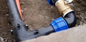 Close-up of black PVC water pipe connected to brass fitting and blue elbow during water line repair service, exposed in excavated dirt trench