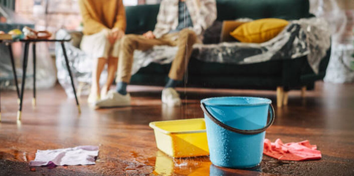 Bucket collecting water leaks from ceiling causing damage to hardwood floor in living room