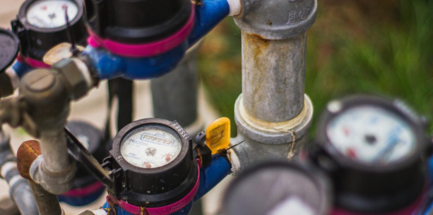 Close-up of a residential water meter assembly and galvanized metal piping outdoors.
