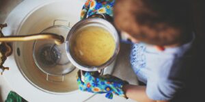 Overhead view of a person wearing colorful oven mitts straining hot pasta water into a metal colander over a white kitchen sink.